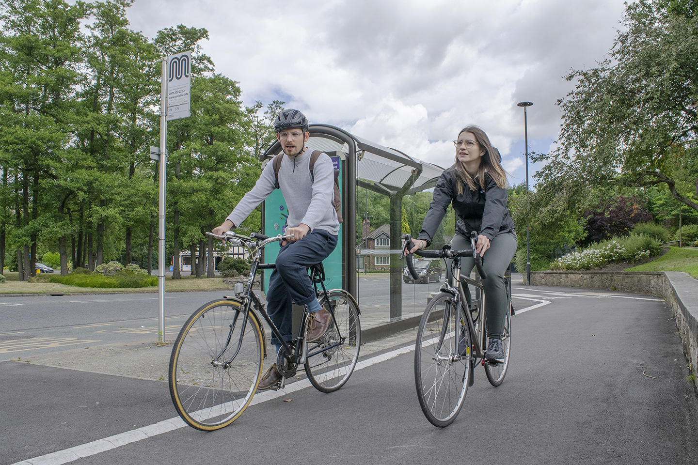 Two people riding bikes on a paved road, near a bus stop.