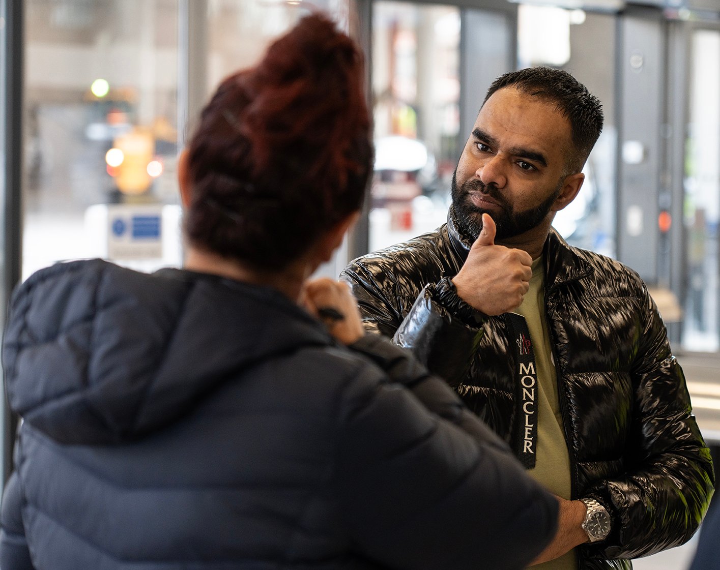 Man in black jacket with thumbs up, Moncler jacket label, woman facing him.