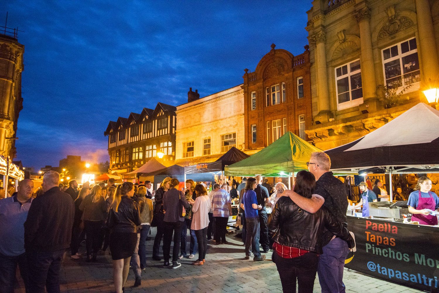 People at a street fair with food stalls and buildings at night.