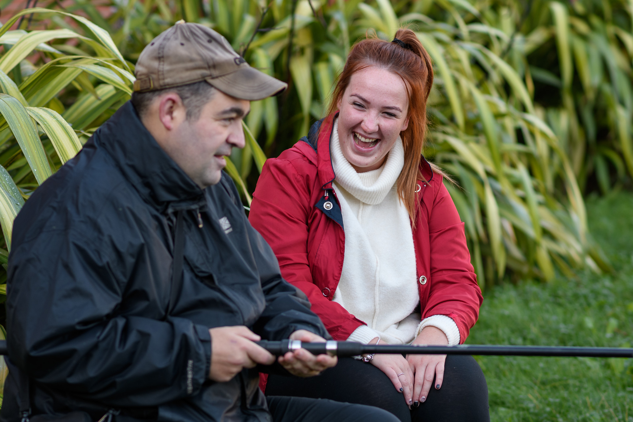 Two people laughing, holding fishing rod, greenery in the background.