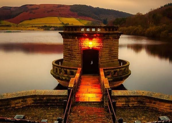 A brick tower and walkway on a lake at sunset, mountains in the background.