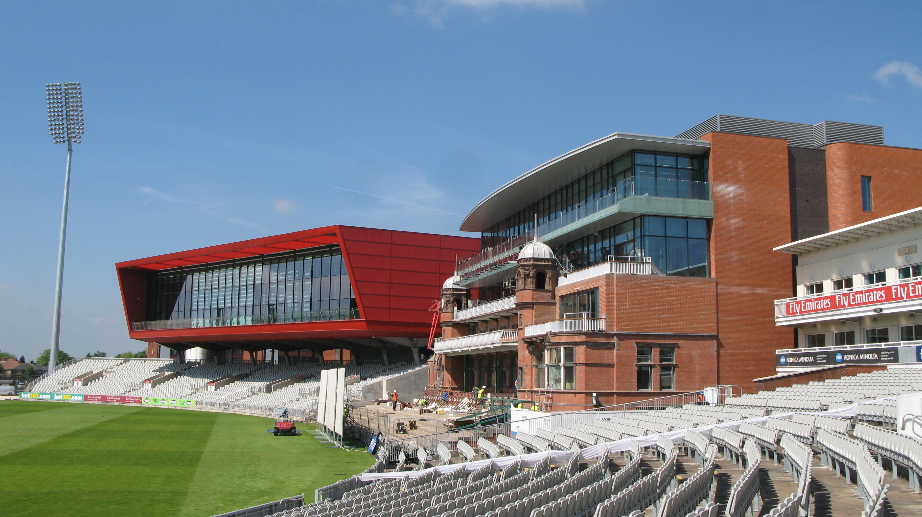 Cricket stadium with rows of seats, green field, and red building.