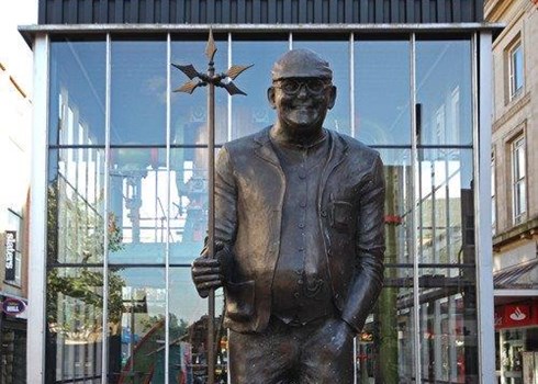 Bronze statue of Fred Dibnah, glasses, cap, holding a prop, in front of glass windows.