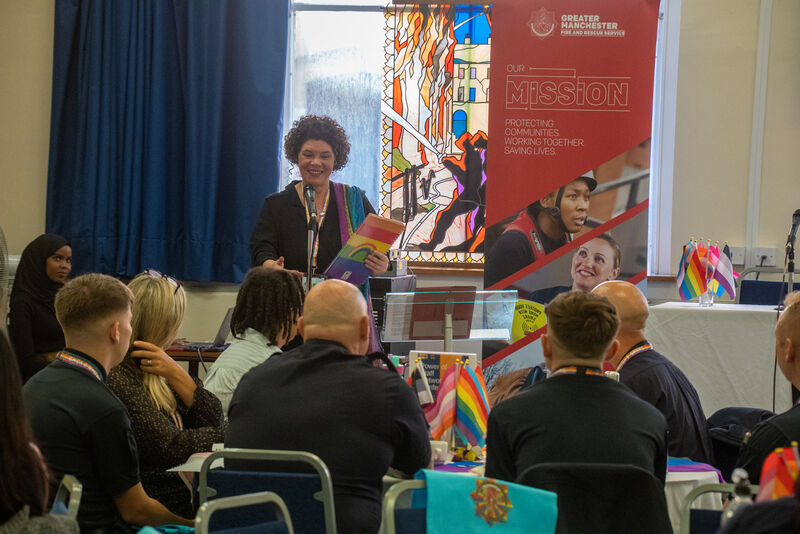 Person at podium speaking with pride flags on table.