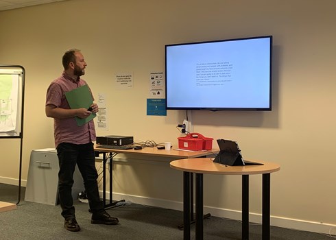 Man giving a presentation; screen with text; table with laptop.