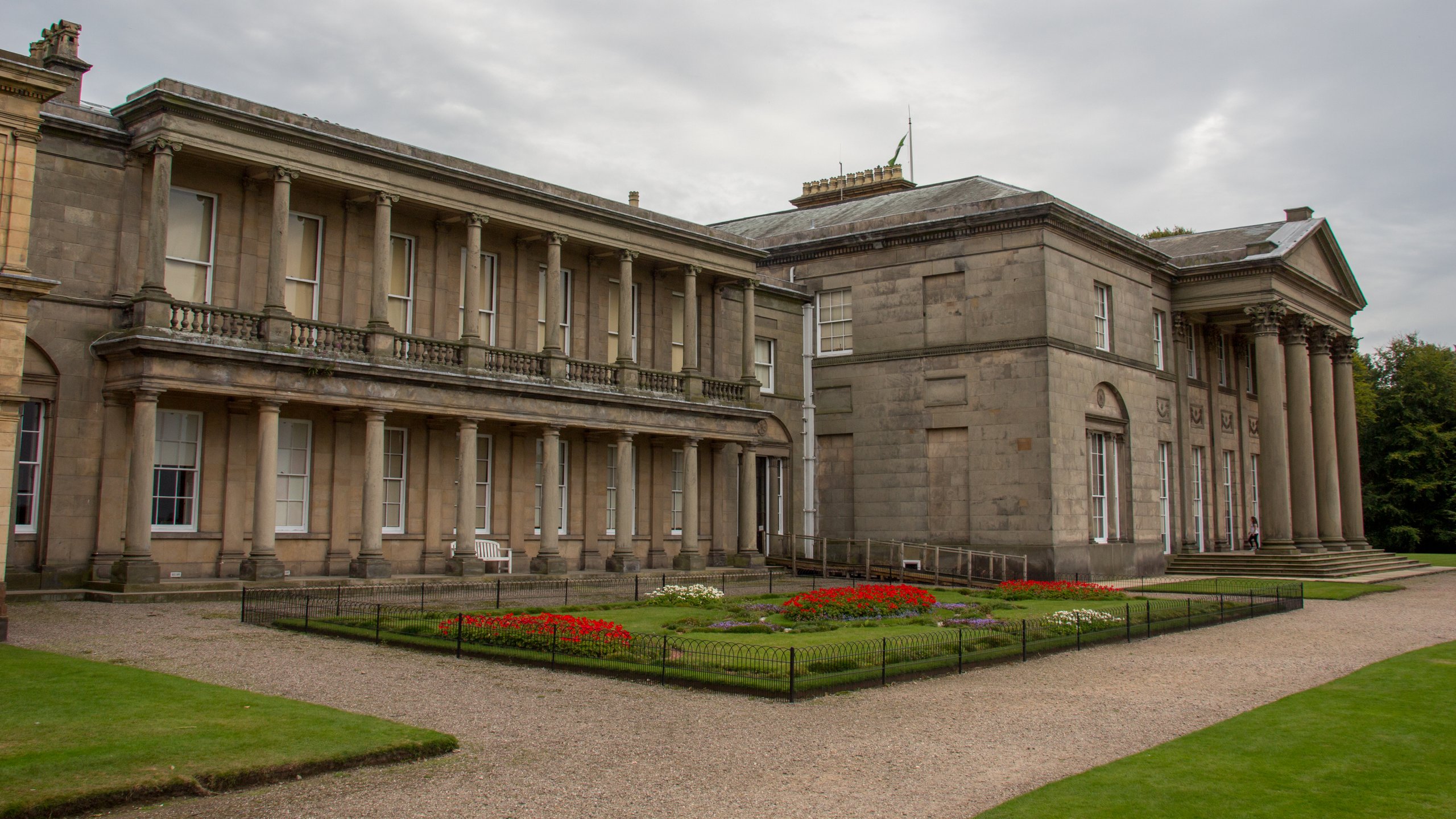 Building with columns, garden with flowers, and cloudy sky.