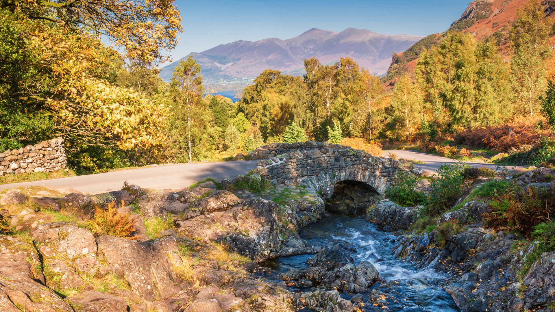 Stone bridge over a stream in autumn scenery. Road, trees, and mountains.