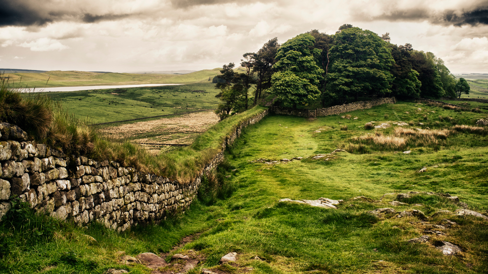 Stone wall in a green landscape with trees and cloudy sky.