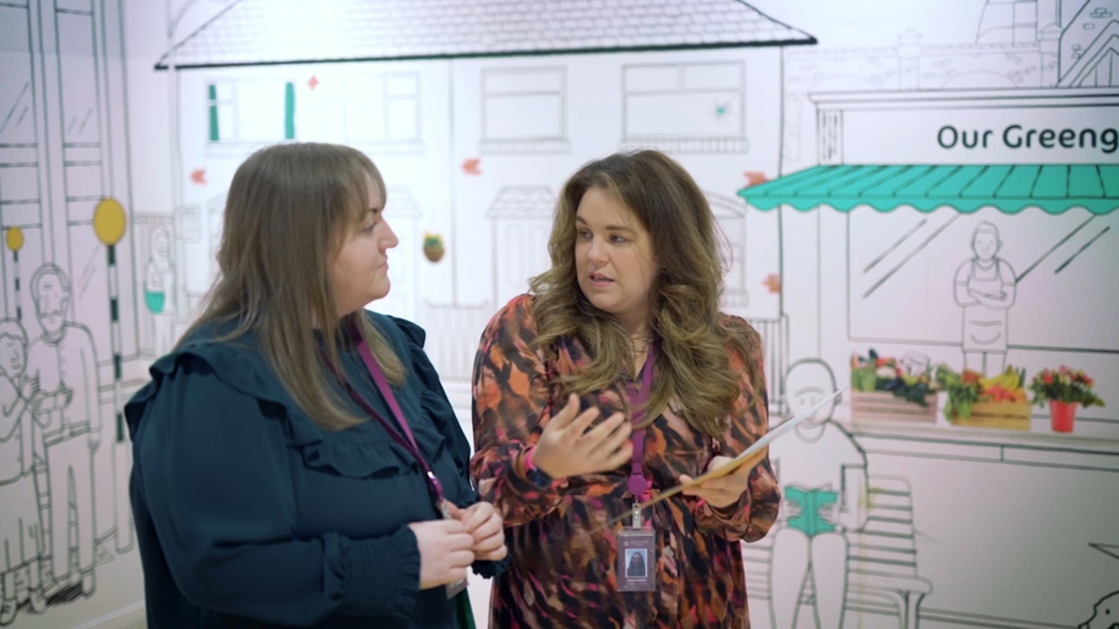 Two women talking in front of a mural with drawn houses and a market scene.