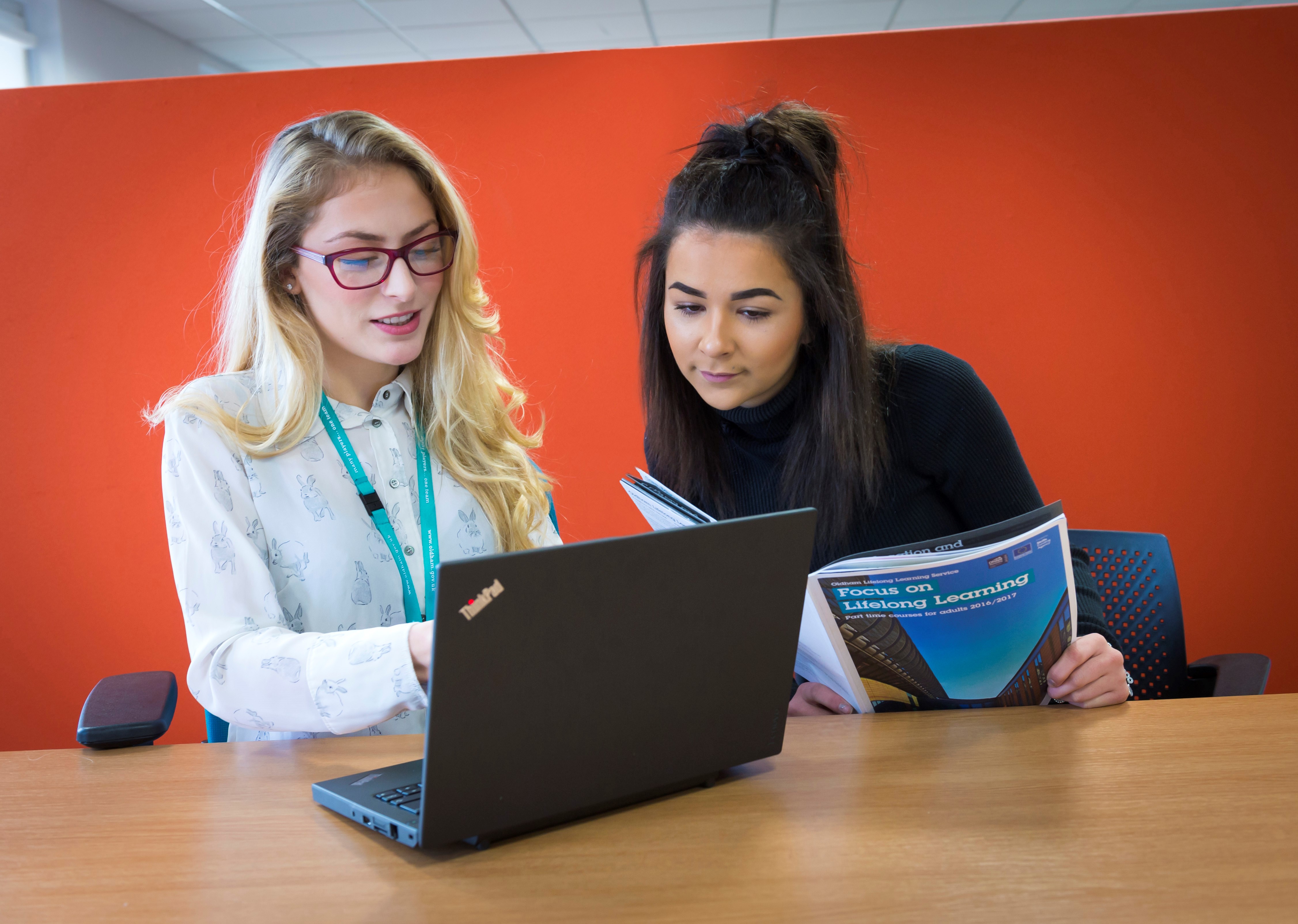 Two women looking at a laptop and a brochure. One has glasses.