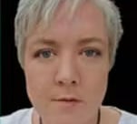 A woman with short grey hair and a white top is looking at the camera.