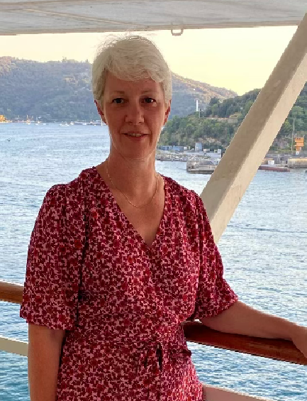 A woman with short white hair wears a red floral dress and stands on a ship's deck. The sea and a hilly coastline are visible in the background.