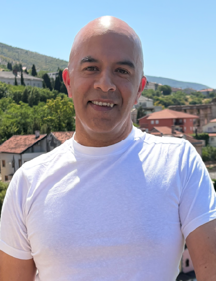A smiling bald man wearing a white t-shirt stands outdoors with a town and hills in the background.