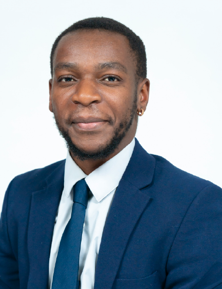 A man wearing a dark blue suit, white shirt, and dark blue tie smiles at the camera. He has a beard and a gold hoop earring.