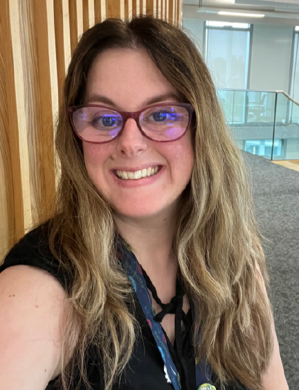 A woman with blonde hair and glasses smiles at the camera. She is wearing a black shirt and a lanyard.
