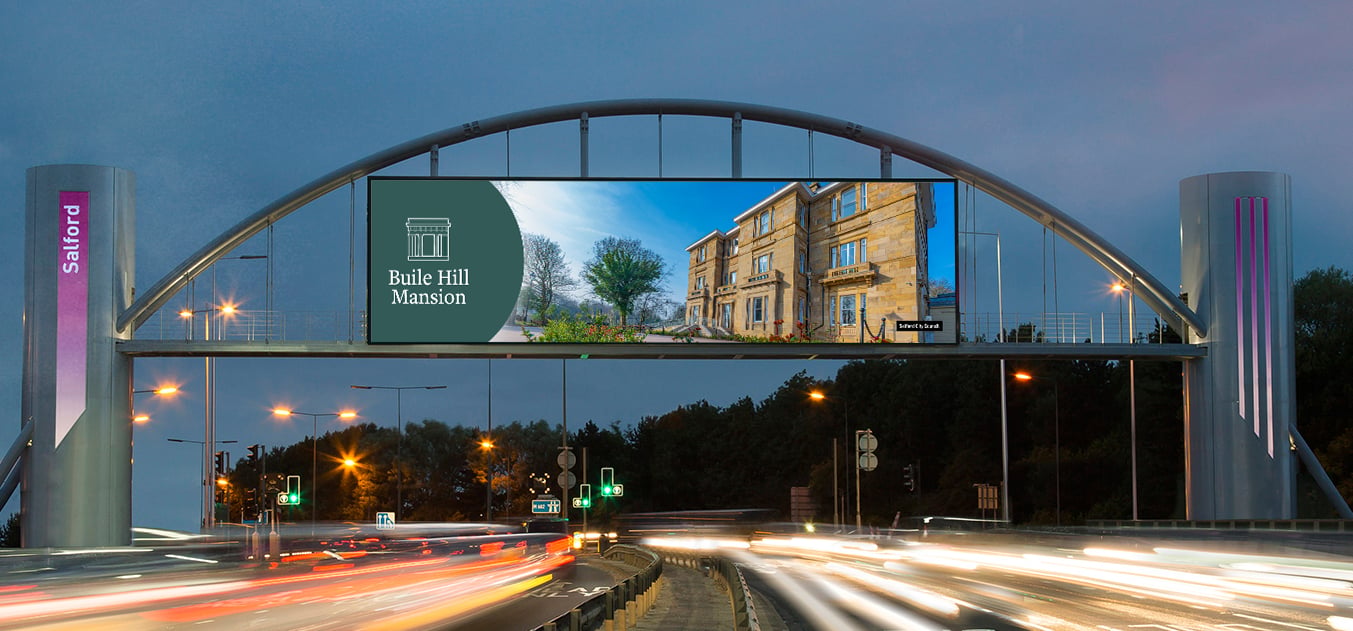 An illuminated archway spans a highway at dusk, displaying an ad for Buile Hill Mansion and a "Salford" sign. Light trails from passing cars blur the road below.