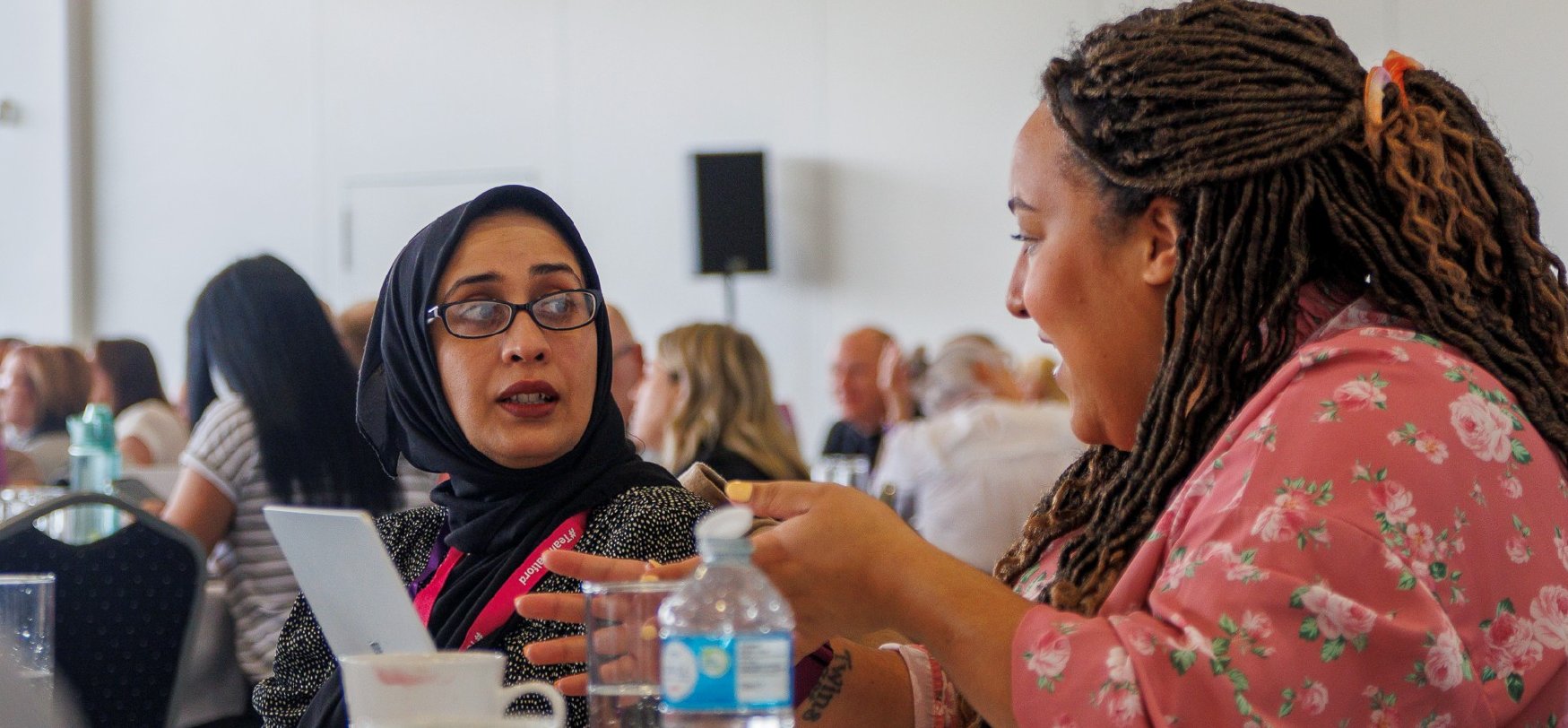 Two women engaged in a professional conversation at a conference table with laptops and drinks.